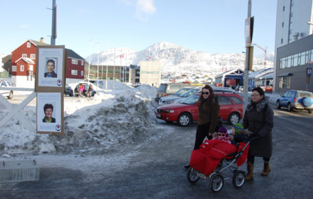 Inuit. Nuuk, at present. Photo of a street in the capital, Nuuk, with sun and snow, election posters, two Inuit women and a baby in a cart.